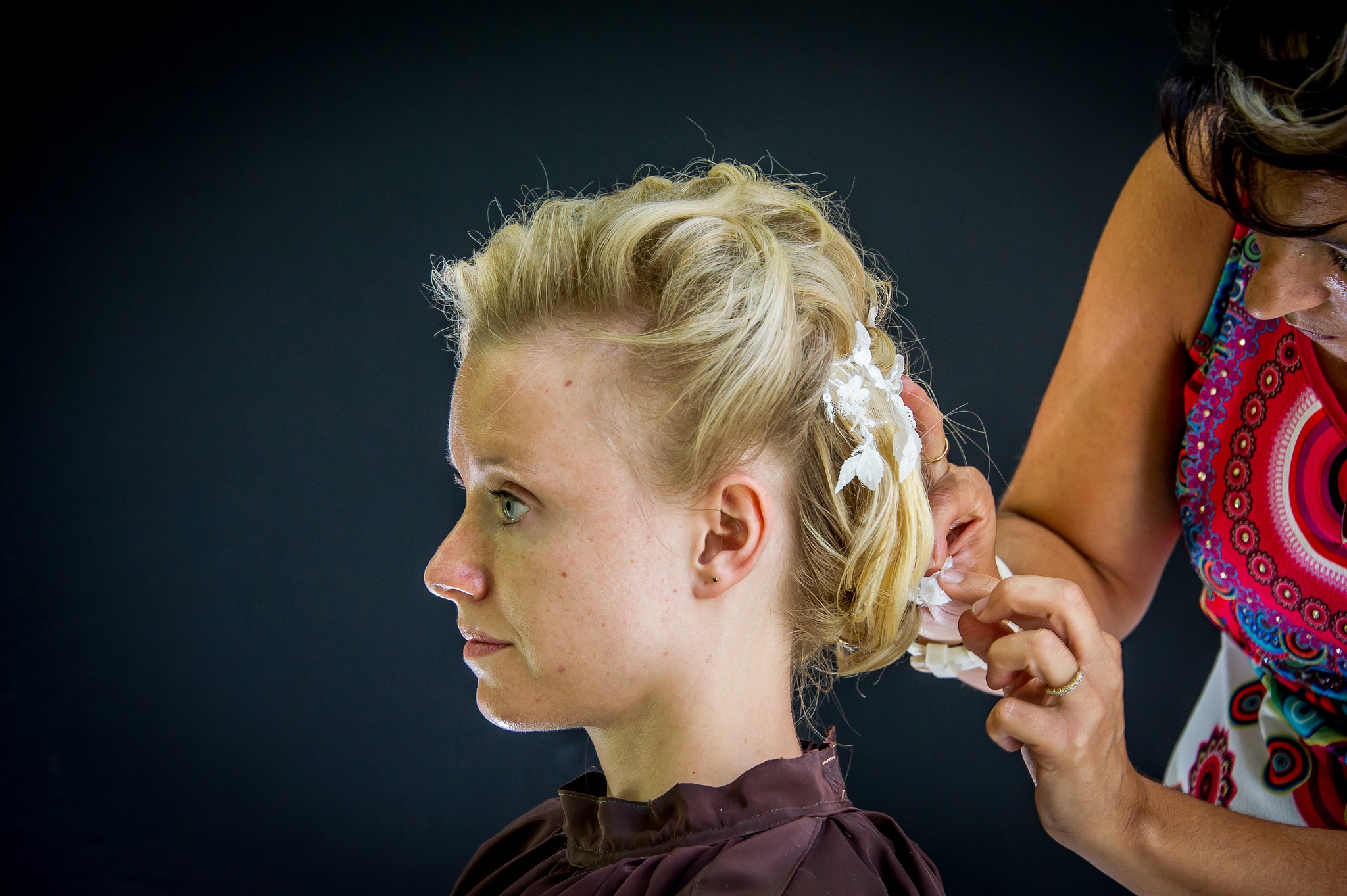 La mariée assise de profil, sa coiffeuse ajustant des fleurs blanches dans ses cheveux