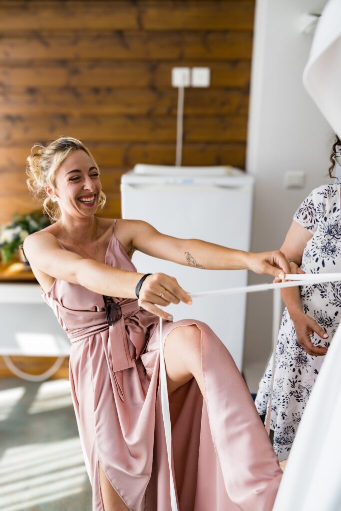 Témoin en robe rose souriante tirant sur les lacets d'une robe de mariée blanche