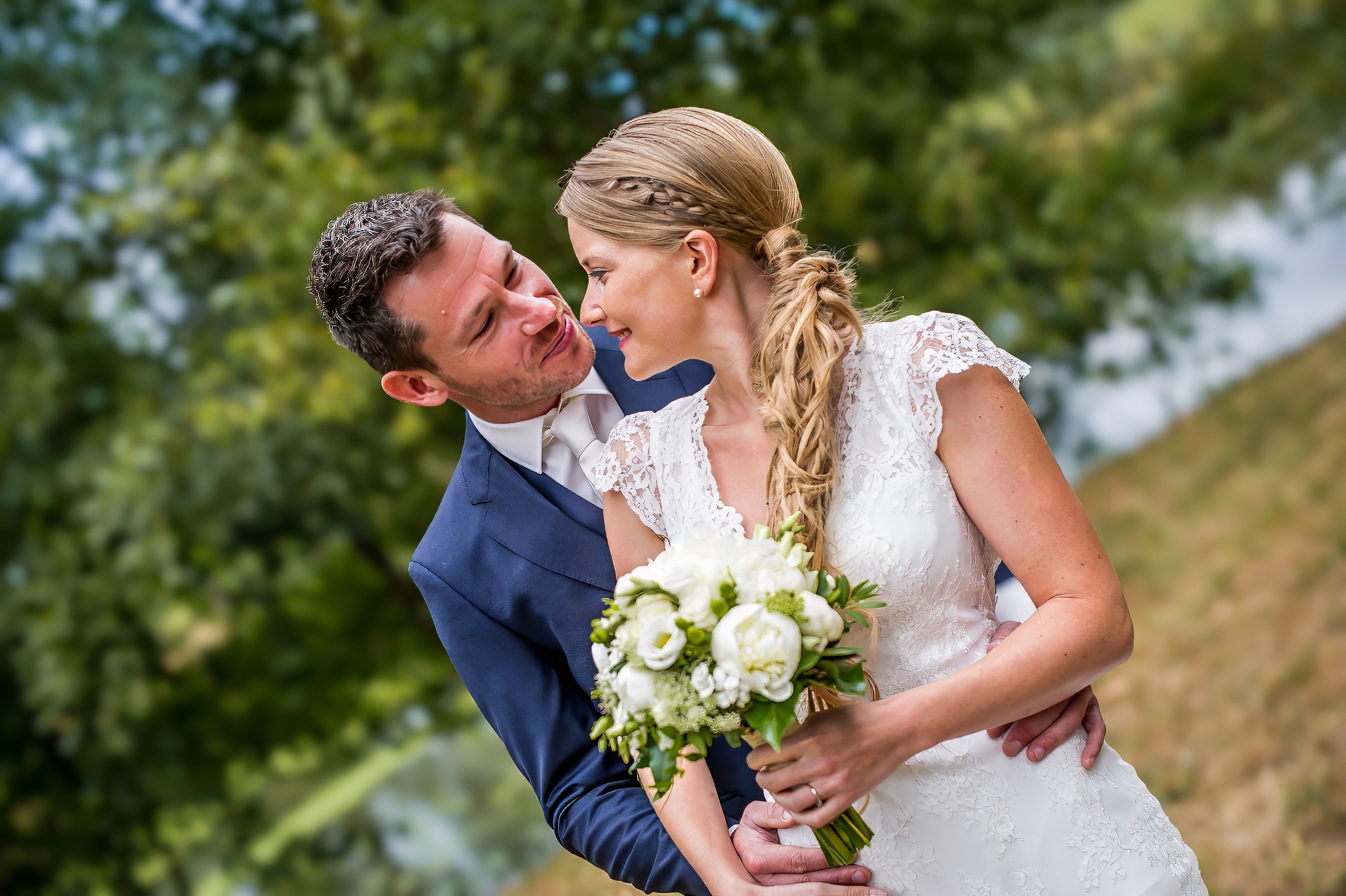 le Marié en costume bleu enlacant sa femme en robe de mariée tenant un bouquet de fleurs blanches, se regardant affectueusement