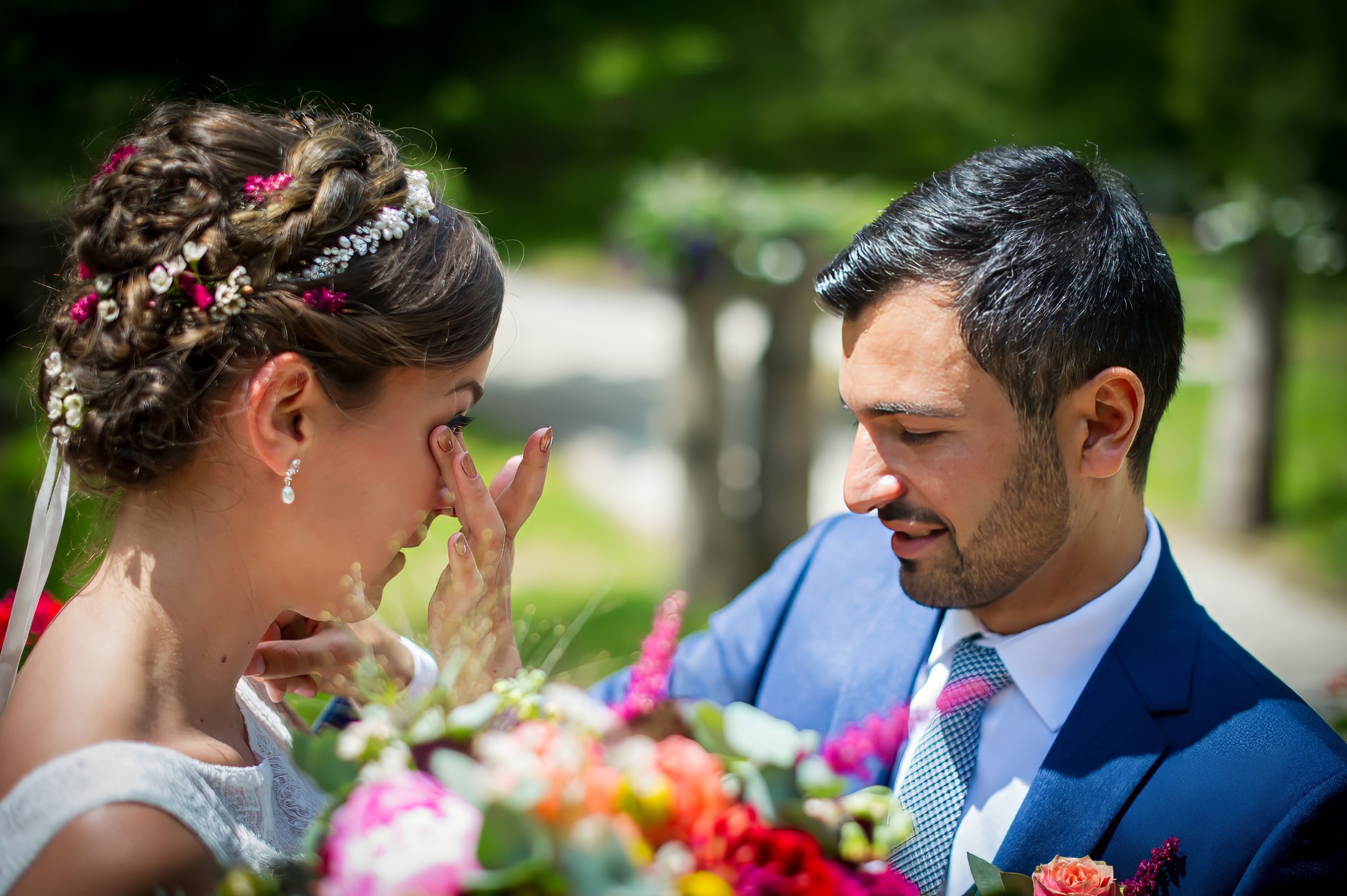 Homme en costume bleu regardant sa femme en robe de mariée essuyant des larmes et tenant un bouquet de fleurs colorées